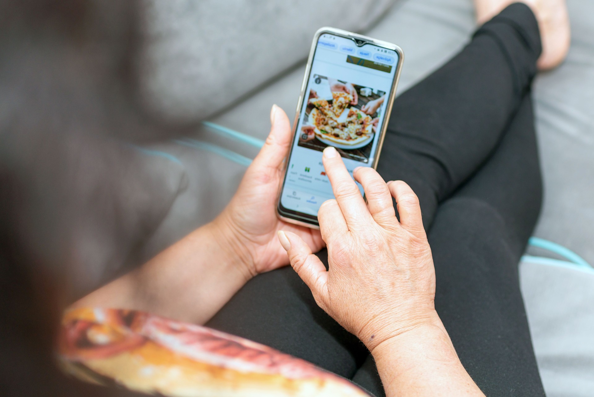Woman holds mobile phone with a pizzeria website on the screen while lying on a fluffy gray couch. Woman orders pizza via phone at home.